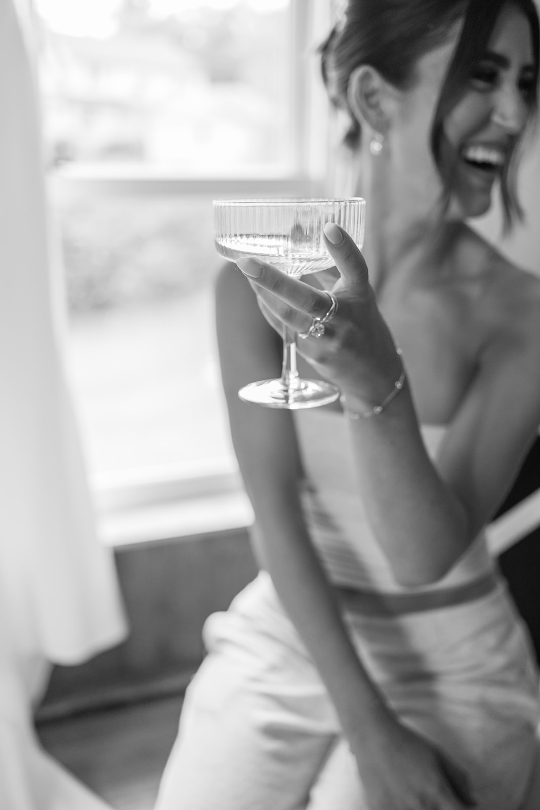 Black and white close-up of the bride holding a champagne coupe, laughing joyfully in the background.