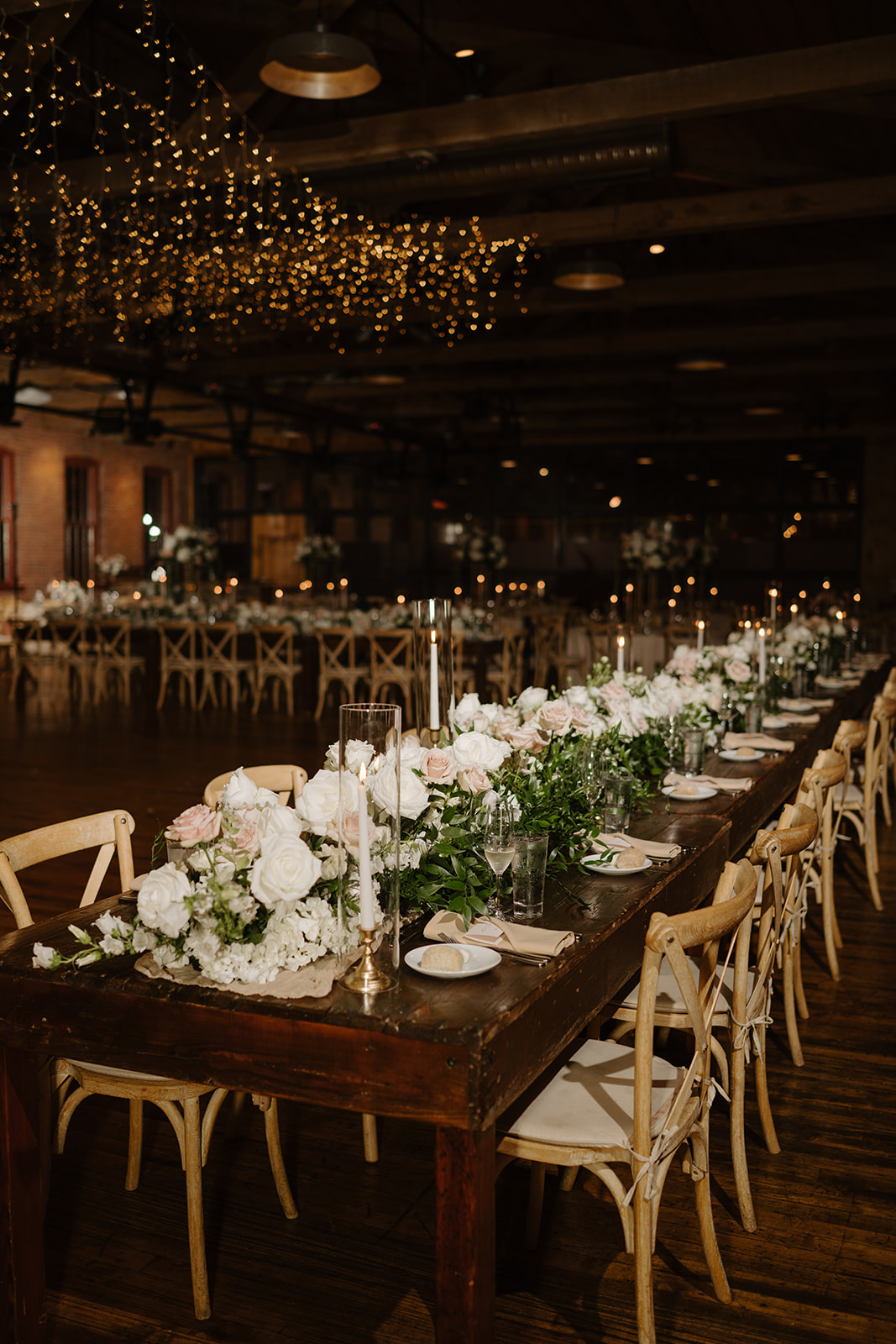 Elegant wedding reception setup featuring long wooden tables, floral runners, and glowing fairy lights.