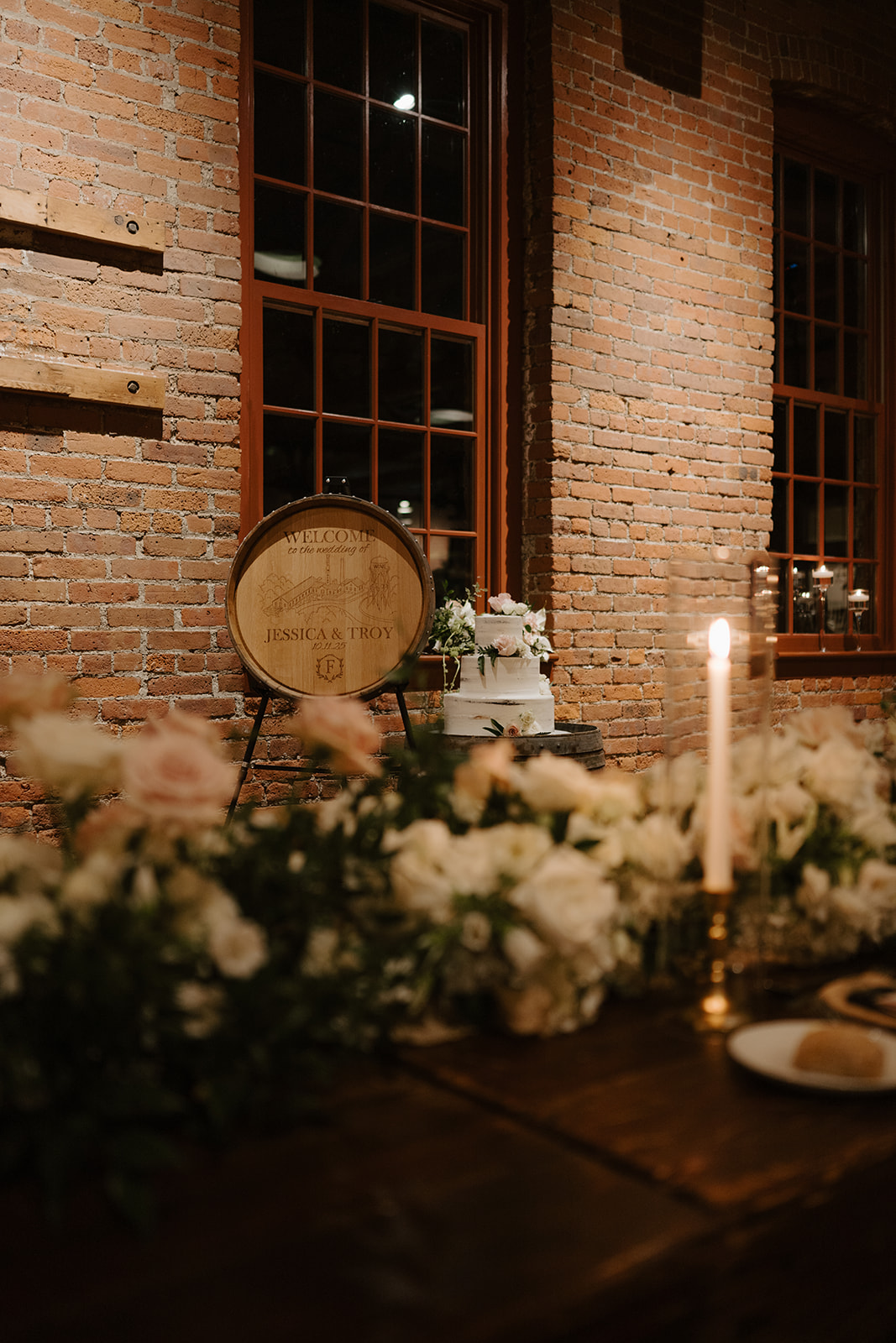 Custom engraved wine barrel lid welcome sign next to a tiered wedding cake by brick walls.