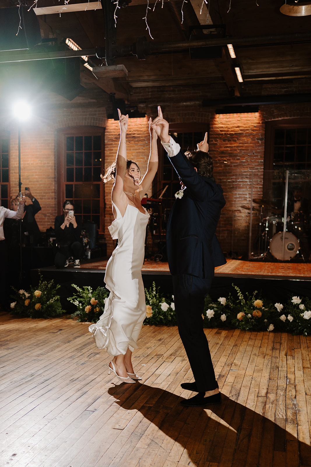 Bride and groom dance enthusiastically with their arms raised high on the wooden floor.