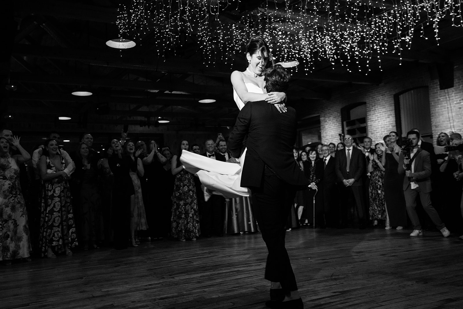 Black and white photo of the groom lifting the bride on the dance floor as guests cheer in the background.