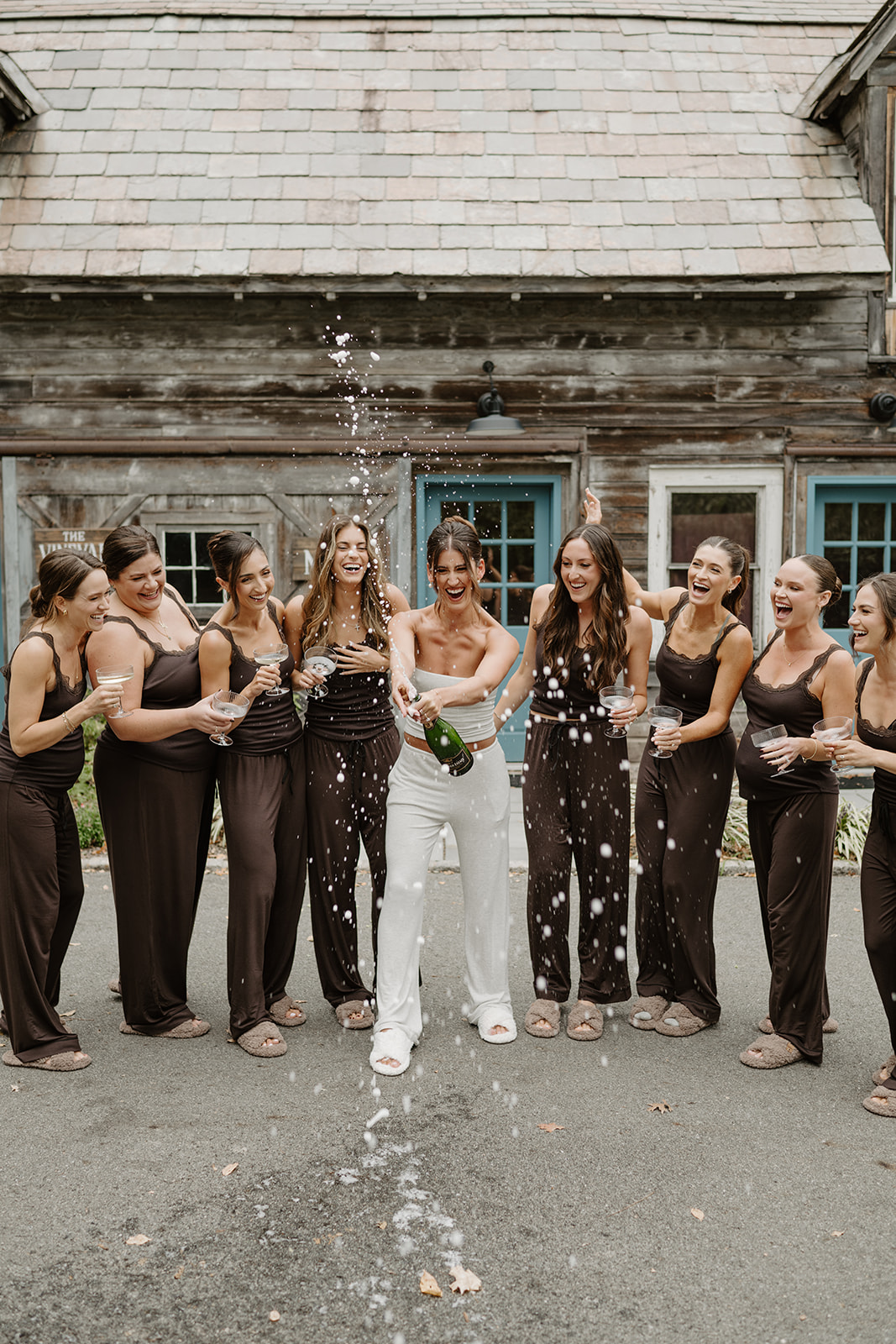 Bride pops a bottle of champagne with cheering bridesmaids in front of a rustic wooden barn.