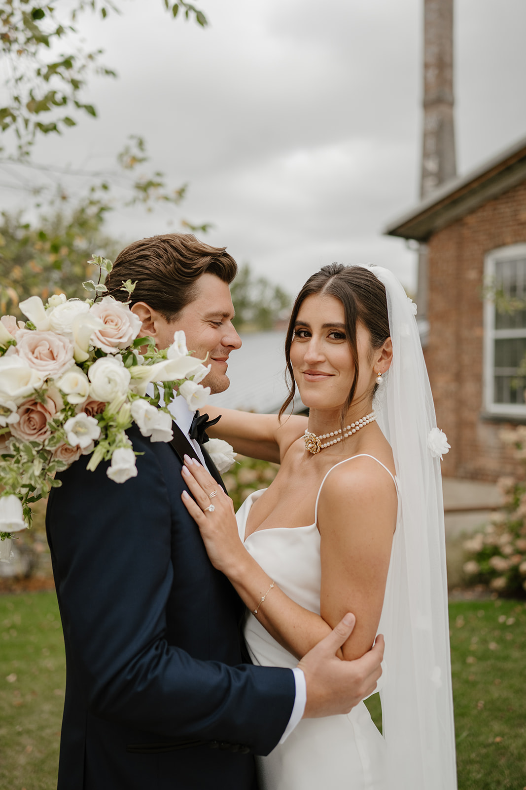 Bride holding a white bouquet smiles at the camera while embracing the groom outside.