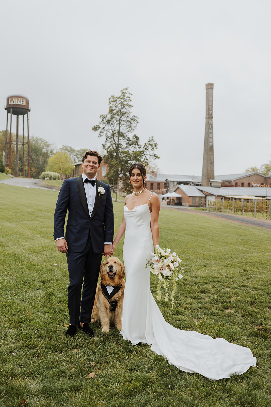 Bride, groom, and their golden retriever pose on a grassy lawn with a wine water tower in the background.