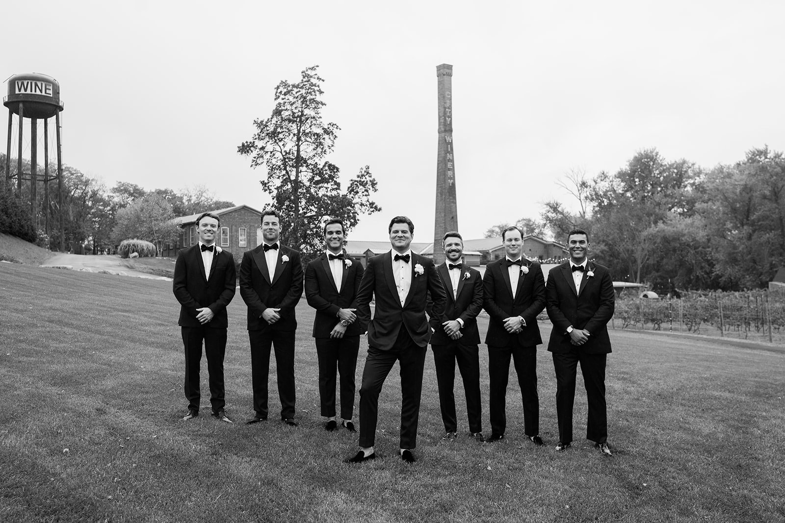 Black and white film portrait of groom and groomsmen posing in tuxedos on a grassy lawn.