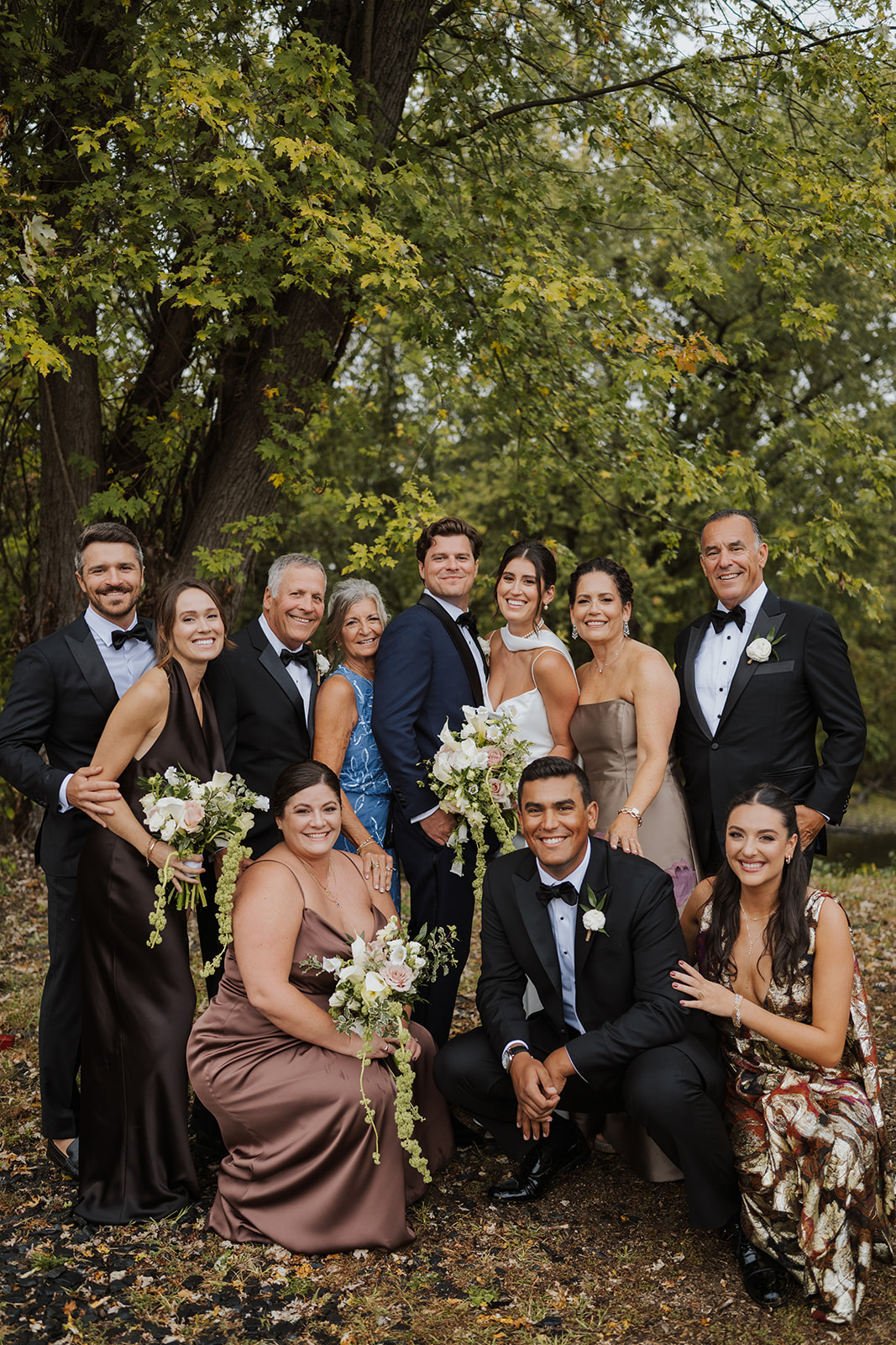 Bride and groom pose for a family portrait outdoors under a canopy of green trees.