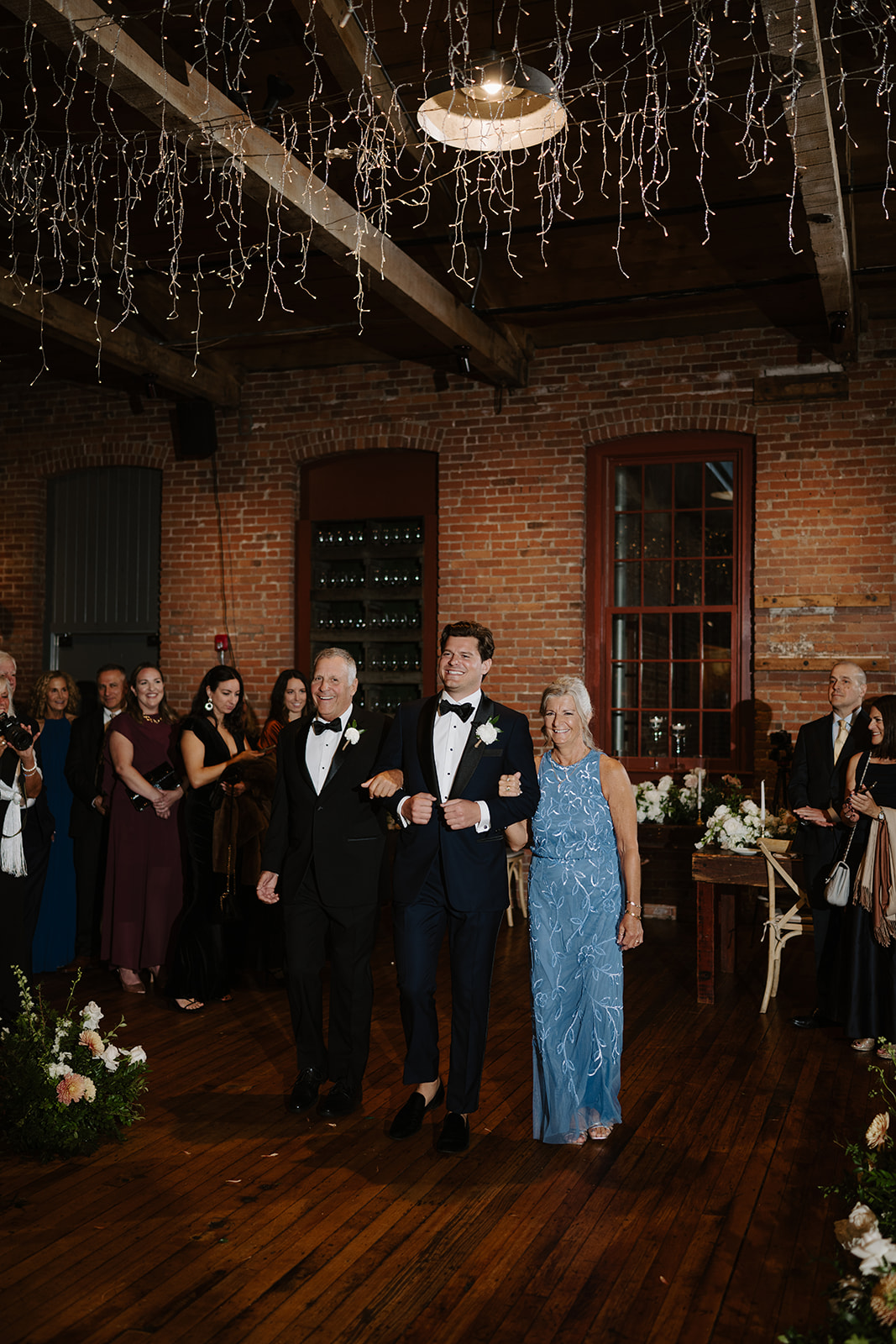 Groom walks arm-in-arm with his parents down the aisle inside a brick-walled venue.