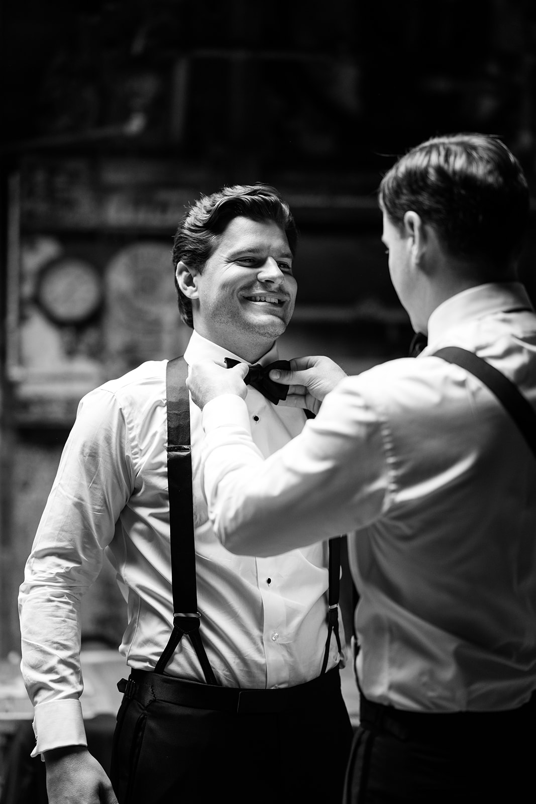 Black and white photo of the groom smiling as his best man adjusts his bow tie.