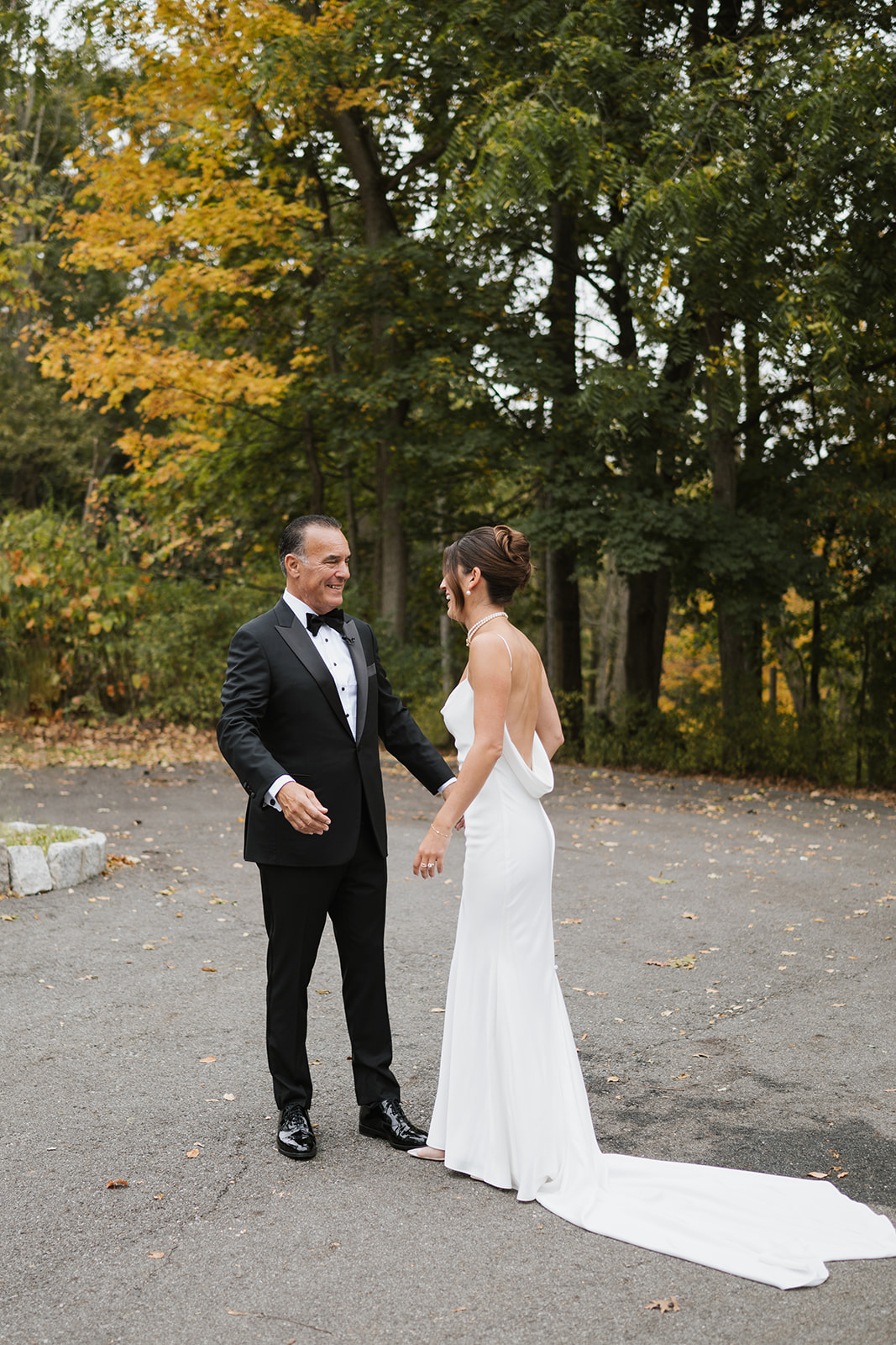 Father and bride smile warmly at each other during their first look in front of autumn foliage.