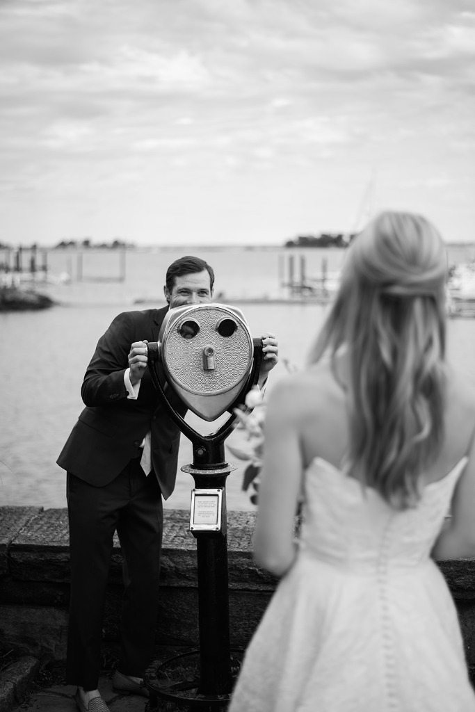 Playful groom looking through shore telescope at bride, editorial black and white wedding portrait